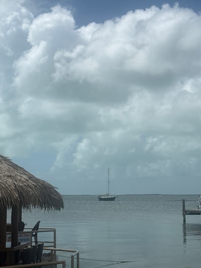 The scene captures a cloudy sky over a body of water, likely in Key Largo, Florida, United States of America. A sailboat is visible on the water. A thatched roof structure and an outdoor seating area are in the foreground.