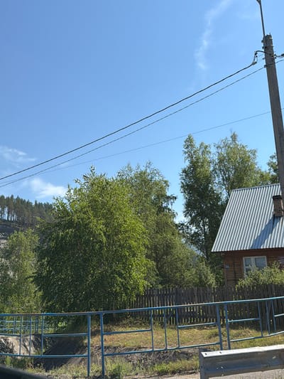 The image depicts a clear blue sky with scattered clouds over a rural landscape. A concrete utility pole with power lines stretches diagonally across the upper right portion of the frame. Below the power lines, a dense collection of deciduous trees with vibrant green foliage dominates the mid-ground. To the right of the trees, a wooden house with a corrugated metal roof and a visible chimney is partially obscured. A rustic wooden fence runs horizontally across the middle of the image, separating the foreground from the background. In the foreground, a blue metal railing runs along what appears to be a pathway or embankment, with a grey metal barrier in the lower right corner. The scene is set in the city of Uznezya, Russia, suggesting a tranquil, natural environment.