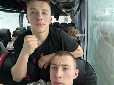 Two fair-skinned teenage boys pose for a photo on a bus. One boy wears a black t-shirt, and the other wears a light-colored shirt. Safety belts are visible. Outside the bus window, raindrops streak the glass. Bus seats and other passengers are visible in the background. The image has been rotated.