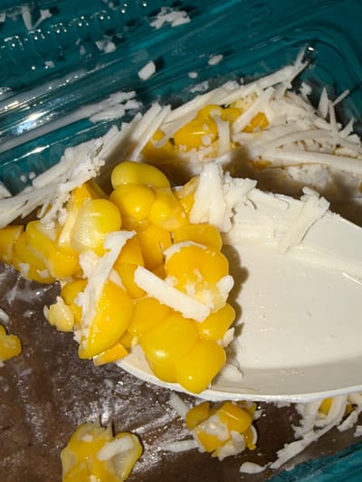 A close-up, overhead shot shows a white plastic spoon filled with corn kernels and shredded coconut. The spoon rests on a brown surface, possibly a table or counter. The corn is bright yellow and appears fresh. The shredded coconut is white and fluffy, mixed in with the corn. In the background, a teal-colored plastic container with a ribbed texture is visible, also containing corn and coconut. The lighting is somewhat dim, with some reflections on the plastic surfaces. The image does not depict any identifiable landmarks or scenes from Tupah, Indonesia.