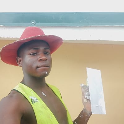 A man in a red hat and yellow tank top is applying white plaster to a wall with a trowel. Paint is splattered on his hand, arm, and tank top. The wall is painted in an unidentifiable color. The scene appears to be set at a construction site or in a partially renovated building. The location is Gana Daji, Nigeria.