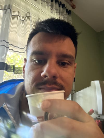 A man is holding a small white cup in his right hand. He is likely indoors, as seen by the curtains and window with a view of greenery and trees, possibly located in Dolni Rakovets, Bulgaria. The room has green walls and a white cabinet. The man has dark hair and a mustache. He is wearing a blue jacket.