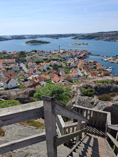 This is an outdoor, eye-level, full shot of a village nestled on the coast of a body of water, as viewed from a wooden staircase. The village consists of numerous houses with orange tiled roofs and white or yellow facades, clustered together on a rocky, sloped terrain. Some of the houses are situated along a street where cars are parked. Beyond the village, the blue water is dotted with small, rocky islands covered in vegetation. A marina with several boats is visible on the right side of the frame. The sky is clear and blue. The foreground is dominated by a wooden staircase and railing, worn and weathered, with some visible graffiti on the railing. The overall impression is of a picturesque coastal settlement on a sunny day. The location is likely Fjällbacka, Sweden, given the characteristic archipelago landscape and architectural style. There are no people visible in the image.