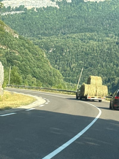 A car trailer carrying three large bales of hay is being towed by a vehicle along a curved asphalt road. The trailer is loaded with two bales stacked horizontally and one bale stacked vertically on top. A tall, thin pole extends from the trailer and leans to the left. The road has white lane markings. The landscape features a steep, densely wooded mountainside on the left, with a lighter-colored cliff face visible at the very top. On the right side of the road, a portion of a dark-colored car is visible. The scene is set in a rural, mountainous environment, likely the region of Lavans-lès-Saint-Claude, France, given the context. The lighting suggests it is daytime. There are no visible people.