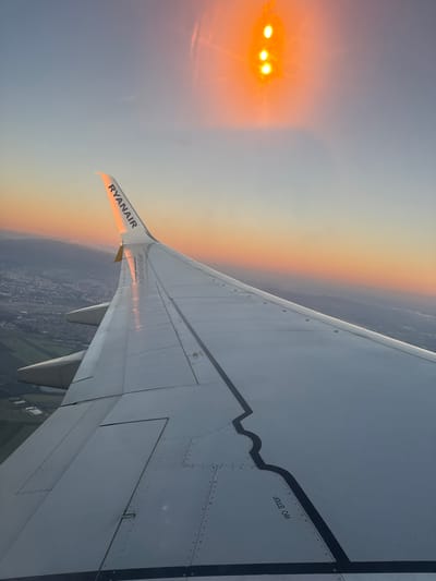 The image shows a view from an airplane window during sunset. The wing of the airplane, emblazoned with "RYANAIR," dominates the foreground. In the distance, a city skyline is visible, with buildings and fields stretching towards a horizon where the sun is setting, casting a warm orange and pink glow across the sky. The sun itself appears as a bright, diffused light source at the top of the frame. The image does not provide any information that would allow for the identification of the city of Hart, United Kingdom.