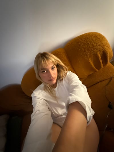 A blonde woman in a white shirt reclines on a large, brown, textured chair. She is facing the camera, looking directly at the viewer. Her right arm is extended towards the camera, partially obscuring the shirt. The background is a plain, light-colored wall. A cable is visible near the top right corner of the frame. The chair appears to be plush and inviting.