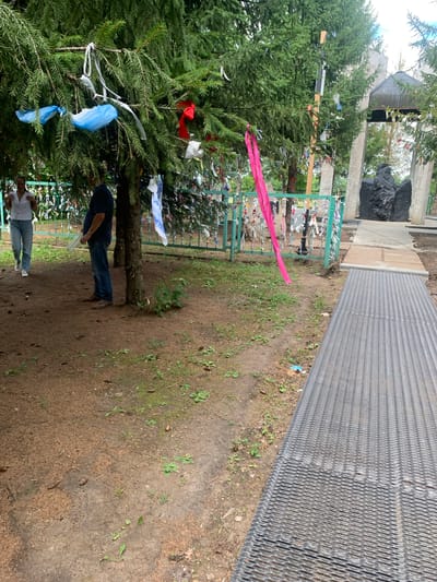 In the city of Arbuzov-Baran, Russia, a scene unfolds around a natural memorial adorned with prayer ribbons tied to trees and a fence. Two individuals are present: a woman in a white shirt and blue jeans walking away from the camera, and a man in a dark blue shirt and jeans standing near a tree, holding a clear plastic bottle. The environment is a wooded area, with lush green fir trees dominating the foreground. The ground is covered in a mixture of dirt, pine needles, and small green plants. Behind the trees, a teal-colored metal fence is visible, also decorated with numerous colorful ribbons. Further in the background, a memorial structure is partially obscured by the trees. It appears to be made of stone pillars and a dark, abstract sculpture. A metal walkway extends from the right side of the frame towards the memorial area.