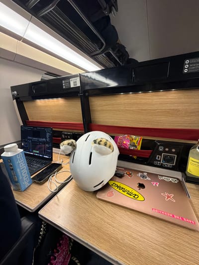 The scene depicts the interior of a modern train carriage, located in or traveling through Bourgoin-Jallieu, France. Two light wood-grain tables are occupied with personal items.
On the left table, a black open-lid laptop displays what appears to be video editing software with a timeline. To its right, a black smartphone rests horizontally, connected by a white cable that extends off-frame. A light blue carton labeled "Pédoures" stands vertically to the left of the laptop. Part of a dark sleeve is visible on the far left, suggesting a seated individual.
Centered between the tables is a white ski or snowboard helmet with two plush, spotted, cat-like figures attached to its sides, resembling ears.
On the right table, a closed rose gold-colored laptop is covered in multiple stickers, including "ammessa," a shell, a starfish, "SAVE THE MERMAIDS," and "La Slesh." Behind this laptop, a yellow bottle labeled "Lemonade" and a yellow bag of "Haribo" gummy candies are visible in a recessed storage area. This area also features a panel with a Type E power outlet and USB charging ports.
The train interior features overhead black panels with integrated LED lighting below and longer fluorescent lights above. Overhead luggage racks are visible above these panels. Red fabric strips line the base of the storage compartments. Text on the overhead panels includes a QR code and "Le Forfait TGV Max."