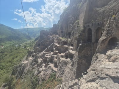 This media file depicts the ancient cave monastery complex of Vardzia, Georgia, nestled within a towering cliff face. The scene is set against a backdrop of lush green valleys and rolling hills under a bright, partly cloudy sky, suggesting a pleasant daytime. A thin cable, possibly from a cable car, stretches across the upper left corner of the frame, hinting at access to the site.

The primary subjects are the intricate cave dwellings and structures carved directly into the rock. Numerous staircases, pathways, and railings are visible, indicating that the site is accessible and likely a tourist attraction. Small groups of people can be seen exploring the various levels of the complex, suggesting active visitation. Notable details include the arched entrances to some of the larger caves, the rough texture of the rock, and the sparse vegetation clinging to the cliff face. The overall impression is one of historical grandeur and natural beauty. The lighting and bright sky suggest it is midday on a clear day. No visible text is present in the media.