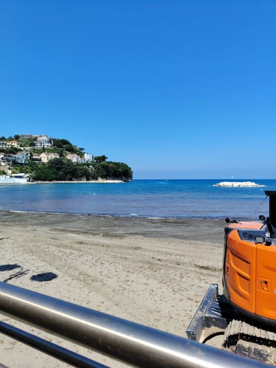 A sunny, clear day at a beach in Agropoli, Italy, shows a wide expanse of sand in the foreground with tire tracks. A section of a shiny metal railing runs along the bottom of the frame. In the mid-ground, the water is calm, with a line of seaweed visible along the shore. A residential area with white and earth-toned buildings is built on a green, tree-covered hill overlooking the bay. Some small white buoys are visible in the water, and a short pier with a white structure on it is in the distance. In the foreground right, an orange construction vehicle with tracks is parked on the sand.