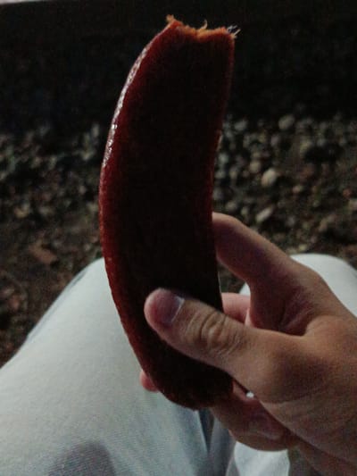 A person's hand is holding a piece of dark red, possibly fruit, that is curved and has a textured surface with some shiny spots. The hand is positioned in the lower right of the frame, with fingers visible. The person is wearing light-colored pants. The background is out of focus and appears to be a dark, possibly outdoor environment with some rocky or gravelly texture. The image appears to be taken at night or in low light conditions, and there is some noise in the image. There is no indication of the location being Novotitarovskaya, Russia.