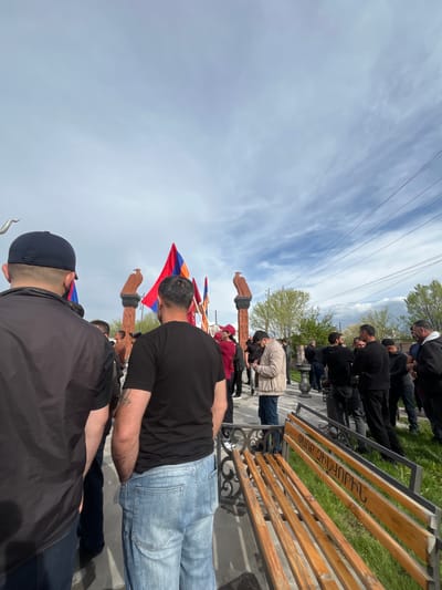 A medium-angle, outdoor photograph depicts a group of people gathered at what appears to be a memorial site in Akhuryan, Armenia. The image is rotated counterclockwise.

The group appears to be paying respects or commemorating an event. They are standing near a series of stone khachkars (Armenian cross-stones), with the Armenian flag visible among them. Several people are wearing black clothing, indicating a possible period of mourning or remembrance.

A park bench with Armenian script on the wooden slats is visible along the top right of the image. A grassy area and trees line the background. The sky is partly cloudy. Power lines are strung across the upper left corner of the frame.