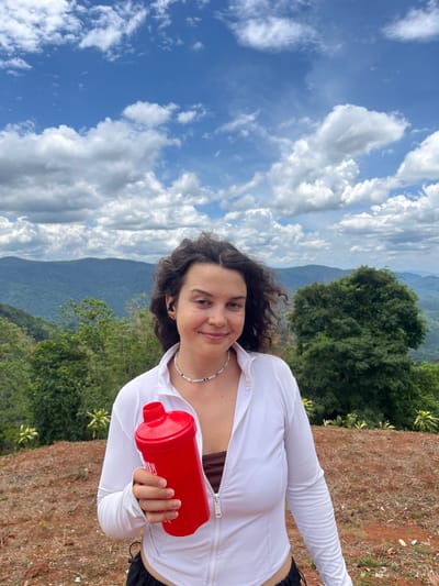 A young woman stands outdoors holding a red shaker bottle in her left hand. She is wearing a white zip-up jacket, a brown top, and black shorts. She has curly dark brown hair and is looking at the camera with a slight smile. The background consists of lush green mountains and a cloudy blue sky. The location is identified as Nao Se, Vietnam. The ground in the foreground is reddish-brown soil.