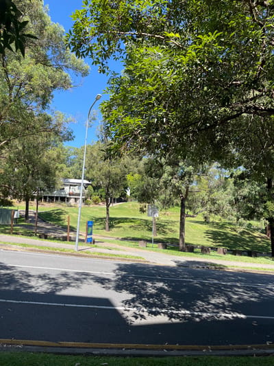 This image, taken in Kelvin Grove, Australia, presents a sunny outdoor scene featuring a lush green park alongside an asphalt road. The primary subjects are the numerous trees, vibrant with green foliage, which heavily populate the left side and background, and cast long, distinct shadows across the park's grass and the road.
The asphalt road, visible on the right, includes a white center line and a yellow line near the curb, bordering a concrete footpath that separates it from the park area. Within the park, several concrete or stone benches are present. A tall street light pole stands prominently, extending from the bottom left towards the center of the frame. Two signs are visible in the park: a larger informational plaque with unreadable text mounted on a post, and a smaller blue and green sign on a shorter pole. A portion of a building, possibly a house or other structure, can be seen nestled among the trees in the distance.
The bright sunlight and pronounced shadows indicate that the time of day is either mid-morning or early afternoon. The weather is clear and sunny, evidenced by the brilliant blue sky visible through the tree canopy. No people or specific activities are depicted, giving the scene a calm and tranquil atmosphere.