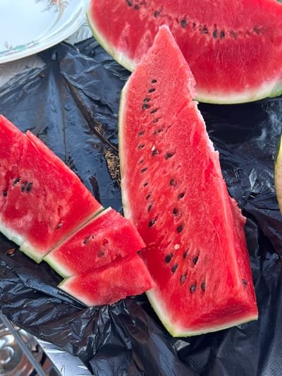 A close-up, overhead shot shows several slices of fresh, red watermelon with black seeds, arranged on a crinkled black plastic surface. To the left, two triangular slices are stacked, one on top of the other, with the top slice appearing to be cut into smaller pieces. A larger, more substantial slice dominates the center and right side of the frame, also showing its red flesh and seeds. In the upper left corner, the edge of a decorative white plate with a floral pattern is visible. The environment appears to be outdoors, possibly at a picnic or gathering in Pribrezhnyi, Russia, suggested by the presence of food and a disposable plastic covering. The lighting is bright, highlighting the vibrant colors of the watermelon.