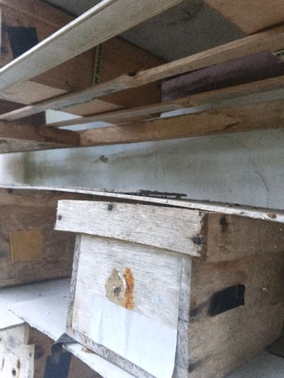 The image presents a close-up, upward-angled view of a structured environment, likely indoors or under a canopy. The upper third of the frame reveals an overhead assembly of aged wooden planks and beams, some exhibiting natural grain variations and others showing lighter, more uniform tones. A segment of a green label, partially reading "MADA", is affixed to one horizontal plank. Beneath this overhead structure, a light-colored wall with visible cracks and minor discolorations forms the background. The lower two-thirds of the image focus on multiple weathered wooden boxes, arranged in a stacked or tiered configuration. The most prominent box, centrally positioned, is constructed from light-colored wooden planks fastened with visible nails. Its front surface features a roughly circular opening surrounded by an irregular, orange-brown residue, below which a rectangular piece of white material is attached. A dark, rectangular opening is cut into the right side of this box. Additional wooden boxes, some displaying yellow or light-colored rectangular patches, are partially visible behind and below the central box. No individuals, explicit actions, or interactions are visible. The specific location within Kon Tum, Vietnam, is not discernible from the visual evidence.