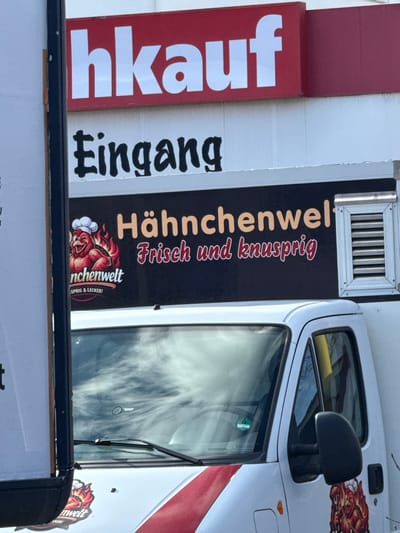 The image depicts an outdoor commercial scene at the Kaufland supermarket on Holländische Straße 135, 34246 Vellmar, Germany.
In the upper portion, a red sign with white capitalized letters, partially visible, displays "...hkauf," indicating a retail establishment. Directly beneath it is a white sign with black stylized text that reads "Eingang" (German for Entrance).
Dominating the lower and central foreground is the front half of a white food truck. The truck's windshield reflects a cloudy sky. A green environmental sticker is affixed to the lower left side of the windshield. A black sign on the side of the truck, positioned above the cab, features orange capitalized text "Hähnchenwelt" (Chicken World). Below this, in red script, are the words "Frisch und knusprig" (Fresh and crispy). To the left of this text, a cartoon logo depicts a red, flame-outlined chicken wearing a white chef's hat. Further down the truck's side, partially obscured by a black vertical post on the left, additional text is visible, including "nchenwelt" and "SPRIG & LECKER!" (Crispy & Delicious!). A silver louvered vent is visible on the upper right side of the truck's black sign. No individuals or specific actions are visible within the frame.