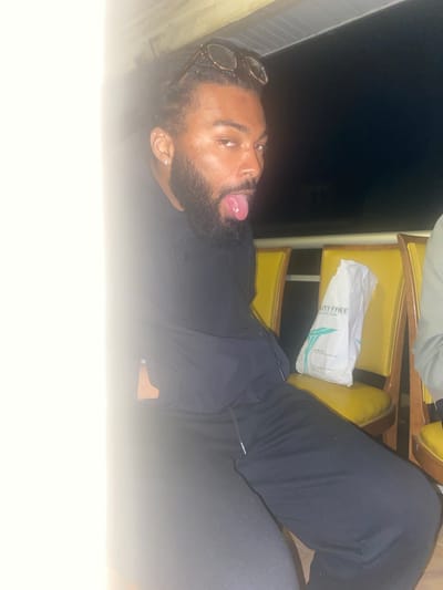 A bearded man with sunglasses on his head is reclining in yellow seats with a duty-free bag beside him, sticking out his tongue. The location is Montreuil, France.