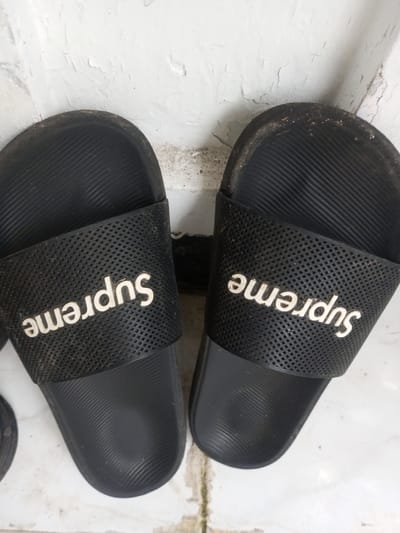 Two black rubber Supreme-branded slides, heavily used, are positioned side-by-side on a dirty surface. The left slide displays "Supreme" in white lettering. The right slide's lettering is inverted. The slides have a ridged footbed and perforated upper band. Dust and grime are visible on the shoes and the surrounding surface. A portion of another shoe is visible on the left. The background features a cracked and stained white wall.