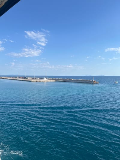 The image depicts a view of a harbor or port facility under a clear blue sky with scattered white clouds. The foreground is dominated by the bright blue, slightly rippled sea. A long, paved pier extends from the right side of the frame into the sea, with a breakwater made of large rocks and concrete tetrapods forming a barrier along its edge. On the pier, several vehicles, including what appear to be trucks, buses, and possibly shipping containers, are visible. Tall lampposts are also spaced along the pier. At the far end of the breakwater, there is a lighthouse or navigational marker. In the distance, across the sea, faint outlines of islands or landmasses can be seen. A small white boat is sailing on the sea to the right of the breakwater. The scene appears to be in the Municipal Unit of Kastro - Kyllini, Greece, given the characteristic landscape of a Greek port.