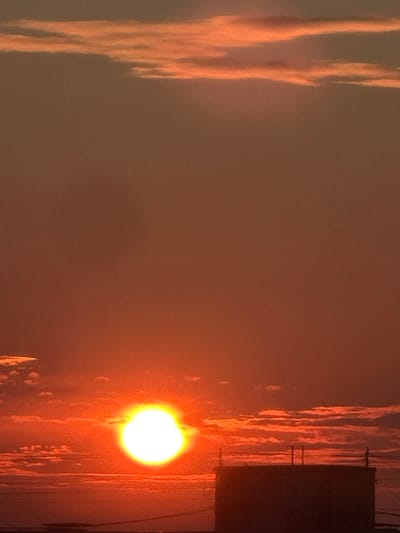The image depicts a sunset over a city, likely Bakhchysarai, Russia, as indicated by the context. The sky is a warm gradient of orange and red, with horizontal streaks of clouds illuminated by the setting sun. A large, bright, spherical sun is prominently visible in the lower left quadrant of the image, emitting a strong glow. In the foreground, the silhouette of a building is visible, appearing dark against the bright sky. This structure has a flat roof and what appear to be antennas or chimney-like protrusions on top. The overall scene suggests a serene end to the day, with the urban landscape partially obscured by the dramatic natural light.