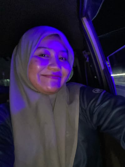 A woman wearing a beige hijab and a dark blue jacket is captured in a selfie from within a vehicle. The lighting is predominantly blue, casting a distinct glow on her face, highlighting her smile. The background is dark, suggesting it is nighttime, with some blurry lights visible through the car window on the right. The location is identified as Sungai Paring, Indonesia.