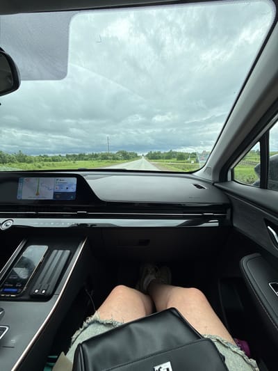 The image, taken from a passenger's perspective, shows the interior of a modern car and the view through its windshield. The sky outside is overcast with visible clouds. A straight road extends into the distance, bordered by green fields or trees and a utility pole with power lines, indicating a rural or semi-rural setting.
Inside the car, the lower body of a person, likely a passenger, is visible on the right, wearing distressed shorts and sneakers, with a black handbag on their lap. The sleek dashboard features a large infotainment screen displaying navigation details, air vents, and various controls. A smartphone is placed in a central console charging area.
Visible text on the screens includes navigation information: "86 км" (86 km) to "ул. Дзержинс" (likely Dzerzhinsky Street), an estimated arrival time of "18:40 У цели 1:51 ч 157 км" (18:40 To destination 1:51 h 157 km). Other details show "HUD Speed," the current time "16:49," a speed of "85.1 км/ч" (85.1 km/h), and a distance of "1114.4 км" (1114.4 km). Air conditioning controls are set to "AUTO AC." The Cyrillic text and the location context suggest the image was taken in Ustinovo, Russia.