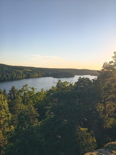 This is a vertical, eye-level, medium-shot photograph of a vast, serene lake surrounded by dense, rolling forest land. The sun is low in the sky, casting a warm, golden light that creates a soft glow on the horizon. The sky is a clear, pale blue, fading to a soft yellow and orange at the horizon. The foreground is dominated by a thick canopy of evergreen and deciduous trees, their branches reaching out towards the viewer. The lake itself is calm and reflects the muted colors of the sky and surrounding foliage. In the distance, the forest continues to undulate, with a few small islands dotting the water. The overall impression is one of natural beauty and tranquility, with the scene likely captured during sunset. The location is identified as Skriketorp, Sweden. There are no people or specific activities depicted in the image.