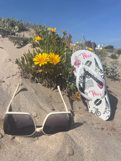 The image depicts a beach scene in La Marina De Creixell, Spain. A white flip-flop with a "Paris" themed design, including depictions of the Eiffel Tower, a lipstick tube, and a bicycle, rests partially on a patch of yellow wildflowers growing in the sand. Nearby, a pair of white-framed sunglasses is partially buried in the sand. The sky is clear blue, suggesting a sunny day. The sand is light brown in color.