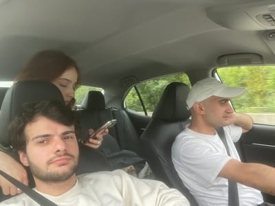 Inside a moving vehicle, likely a car, three people are present. Two males are visible in the front seats, both wearing seatbelts. The driver is wearing a white t-shirt and a white baseball cap. The passenger is wearing a light-colored long-sleeved shirt and holding a phone. The driver is not wearing any visible ring. A third person, possibly female, with red hair, is in the backseat. The environment visible through the car window suggests a wooded or rural area. The image does not suggest any location within Changilari, Georgia.