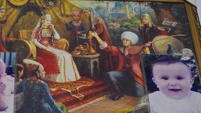 The image is a framed painting that appears to be of a historical or traditional scene, possibly in Kaka, Turkmenistan. The central subject is a woman adorned in regal attire, seated on an ornate chair. She wears a jeweled headdress and elaborate clothing, with intricate patterns on her sleeves and a long, red outer garment over a white dress. Several other figures are depicted around her. To the left, a person in a blue and gold patterned robe is seated, facing away from the viewer, and another figure with a turban stands behind them. To the right, a person in a red jacket and white fur hat is offering a drink from a tray to a woman in a rust-colored dress and head covering. Another figure in a helmet and armor stands behind them, holding a spear. In the background, there are hints of architecture, possibly domes and minarets, and lush greenery, suggesting an outdoor or garden setting. A peacock is visible near the right side of the painting. A small, separate photograph of a smiling baby is placed in the foreground, overlapping the painting slightly at the bottom. The painting seems to depict a moment of ceremony or social gathering. The lighting in the painting suggests daytime. There is no visible text in the image.