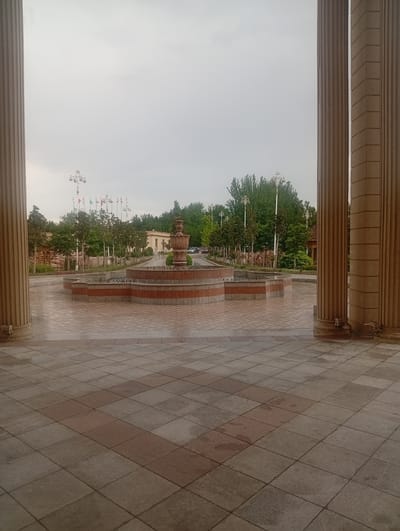 The image depicts an outdoor scene with two large, fluted columns in the foreground, framing a view of a courtyard with a fountain and a pathway. The ground in the foreground is paved with large square tiles, some of which appear wet, suggesting recent rain or mist.
Beyond the columns, a star-shaped fountain with tiered levels and a decorative top is visible. The fountain is surrounded by a low wall. A pathway leads away from the fountain, lined with trees and lampposts. A row of flags from various nations is planted along the pathway, indicating a possible diplomatic or international venue. In the distance, a building with a light-colored facade can be seen, and a few cars are parked or driving on a road.
The overall atmosphere of the scene is calm and somewhat subdued, possibly due to the overcast sky. The time of day appears to be late afternoon or early evening, given the soft, diffused light. The weather seems to be cloudy or possibly drizzling. The location, Guliston, Tajikistan, suggests a formal or public space, perhaps a government building, park, or cultural center, given the presence of national flags. There are no people visible in the image, contributing to the serene and quiet ambiance. The architecture of the columns and the fountain suggests a formal and well-maintained environment.