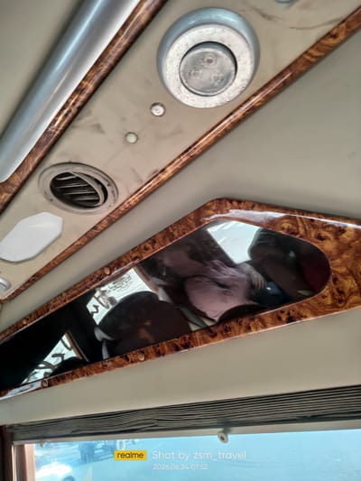 The image depicts the interior of a vehicle, likely a van or bus, with a focus on the ceiling and upper interior trim. The ceiling is upholstered in a light beige material, and a decorative trim with a wood-grain finish runs along the upper edges. Integrated into the ceiling are a circular air vent and a circular light fixture containing three small LED bulbs. A rectangular mirror, framed by the same wood-grain trim, is mounted on the side of the interior, reflecting parts of the cabin, including what appears to be a person's head and shoulder wearing a pink garment. Below the mirror, a roller blind is visible, partially drawn over a window, through which a bright, hazy outdoor scene is discernible, suggesting it might be daytime. The bottom right corner of the image contains a "realme" logo and text indicating it was "Shot by zsm_travel" on "2024.06.24 07:52". The timestamp and the "realme" branding suggest the photo was taken with a mobile device. The location is specified as Gò Dầu, Vietnam.
