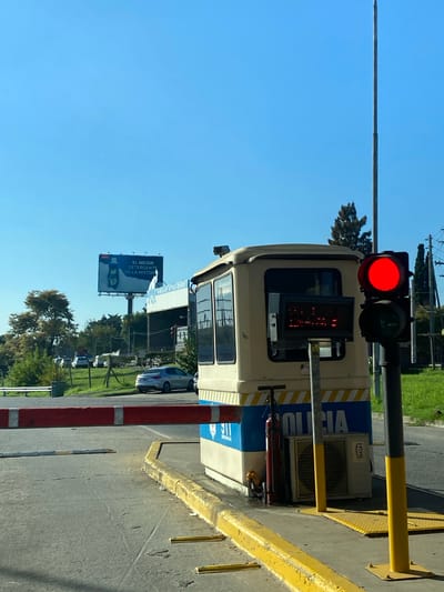 The image depicts a street scene in Don Torcuato, Argentina. A traffic light is visible, showing a red light. Attached to the light fixture is a bus. A billboard is visible in the background featuring an advertisement for detergent. Trees line the street, along with other parked vehicles. The sky is clear and blue.