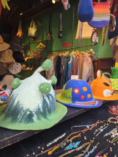 This is a close-up, vertical shot of a market stall selling a variety of handmade felt crafts and jewelry. The primary subjects are colorful felt toys, including a rainbow-striped item with pom-poms, a blue owl-like toy with a circular pattern, and an orange cat-like toy with black eyes and white accents. A large, pale green felt creature with four green, polka-dotted appendages is prominently featured in the foreground, appearing to be a snail or a similar whimsical animal.

The background reveals shelves filled with other felt items, possibly hats and bags, in various patterns and colors. To the right, a display of beaded jewelry, primarily bracelets and necklaces, is laid out on a black velvet surface. The jewelry features a mix of beads, stones, and small bells, with intricate designs.

The setting appears to be an indoor market, possibly a craft fair or a shop, given the arrangement of goods. The lighting suggests it could be daytime, with artificial lights also contributing to the illumination. There are no people visible in the frame. The overall impression is one of a vibrant, creative, and eclectic marketplace. No discernible text is visible in the image.