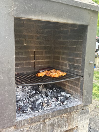 An outdoor brick barbecue grill is shown, with a metal grate positioned above a bed of hot coals. On the grate are several pieces of food, including what appears to be a long sausage and several pieces of chicken or pork ribs. The grill is made of dark, rough-textured brick with a concrete base. In the background, there is green grass and some trees, suggesting an outdoor setting. To the right of the grill, partially visible, are some people and a table, indicating a gathering or event. The location is identified as Campezo / Kanpezu, Spain.