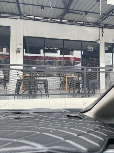 The image shows the exterior of a coffee shop named "Intisari Kopi" located in Blangmetimu, Indonesia. The coffee shop has large glass windows and a metal railing in front of it. Inside the shop, tables and chairs are visible. A sign on the window reads "ORDER & PAY HERE." The scene is viewed from inside a vehicle, with the dashboard in the foreground.