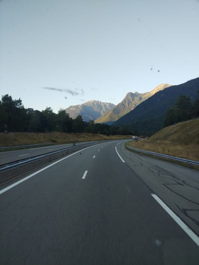 The image shows a highway in the mountainous region near Saint-Léger, France. The highway curves to the right, with two lanes of traffic visible. Guardrails line the sides of the road, and dry grass covers the embankment. In the background, a dense forest of green trees leads up to steep, rocky mountains. The sky is a clear, pale blue, with a few faint clouds. Two trucks are driving on the highway in the distance. The scene appears to be captured from a moving vehicle, as indicated by the motion blur on the road surface.