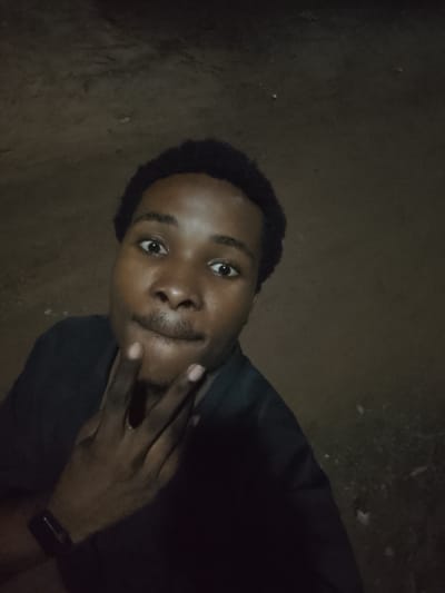 This is a close-up, selfie-style photograph of a young Black person in Orodo, Nigeria. The subject has dark skin, short, dark, curly hair, and is looking directly at the camera with a slightly pensive expression. Their right hand is raised, with fingers touching their chin and mouth area, a common pose suggesting thought or contemplation. They are wearing a dark-colored shirt. The background is out of focus and appears to be an outdoor, unpaved surface, possibly dirt or gravel, with some sparse vegetation or debris visible. The lighting is dim, suggesting it is either nighttime or a very dark, overcast day. There are no visible signs of text or specific landmarks.