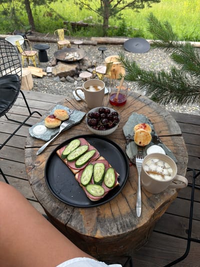 A rustic outdoor setting, possibly a campsite or a lodge in or near the city of Mansky, Russia, features a wooden table laden with breakfast items. The table, appearing to be a large tree stump, is set with a black plate holding a sandwich topped with ham and sliced cucumbers, a small bowl of cherries, and two gray slate plates each with two small pastries and a dollop of cream. A fork rests on one slate plate, and another fork is near a mug of coffee. A glass with a spoon and jam sits beside the cherries. In the background, a fire pit area with rocks and chairs is visible, surrounded by greenery and trees. A person's legs are visible in the foreground, suggesting they are seated at the table.
