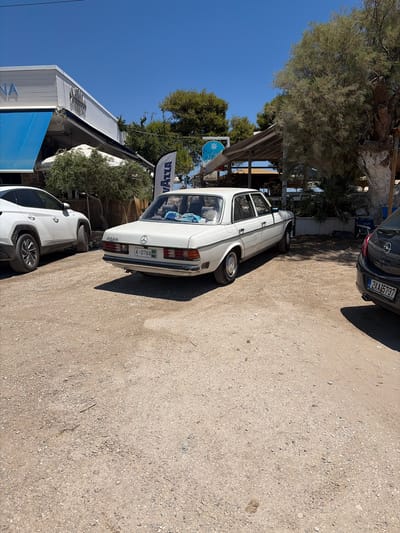 The image captures a daytime scene on a sunny day in Artemida Municipal Unit, Greece. The main subjects are two cars parked on a gravelly surface. The most prominent car is a vintage white Mercedes-Benz sedan. Its rear is facing the viewer, and the license plate, which reads "A-0768", is clearly visible. The car has a light-colored interior and a towel is draped over the back seat. To the left and slightly in front of the Mercedes is a newer white SUV.
In the background, there are trees, some of which appear to be olive trees, and structures that suggest a commercial area, possibly a restaurant or shop. One visible sign has the word "VAZZA" in large, bold lettering. The clear blue sky indicates fair weather. The overall atmosphere appears relaxed and casual, typical of a sunny day in a Greek coastal area. The lighting suggests it is late morning or early afternoon due to the sharp shadows cast by the cars. There is also another car, a dark-colored hatchback, parked further back in the scene, partially obscured by the Mercedes.