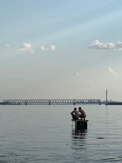 The image depicts a waterfront scene in Пальники, Russia. Two shirtless individuals are on a platform in the water. A lattice-structure bridge spans the water in the background. The sky is light with scattered clouds. The water surface is reflective.