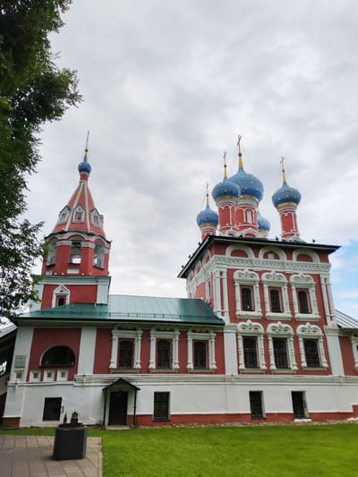 A bright red and white Orthodox church with blue domes topped with golden crosses stands under a cloudy sky. The church has multiple stories, with arched windows and decorative white trim. To the left of the main building is a tall, slender red bell tower with white trim and several openings for bells. A dark green tree with lush foliage is on the far left of the image, partially obscuring the sky. In the foreground, a patch of green grass is visible, with a paved walkway leading to a dark gray, rectangular pedestal holding a small model of buildings. The setting appears to be outdoors, with a focus on the architecture of the religious building. The location is the city of Selivanovo, Russia.