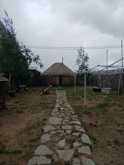 The image shows an outdoor scene in Novoye, Russia. In the center of the frame is a yurt, a traditional portable dwelling. A stone path leads to the entrance of the yurt. To the left of the yurt, there are trees and a small table with chairs. To the right of the yurt, there is a metal canopy structure and a wooden fence made of thin logs. The ground is covered with grass and dirt. A red ball is visible on the ground to the right of the path. The sky is overcast.