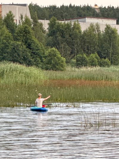 In Glebychevo, Russia, on a body of water, a person is seated in a blue kayak. The individual is wearing a light-colored t-shirt and a hat, with their right arm extended. The kayak is positioned in front of a dense, tall grass shoreline, which is bordered by a line of trees. In the background, there are two large, light-colored industrial buildings visible above the trees. The water is rippled, reflecting the overcast sky.