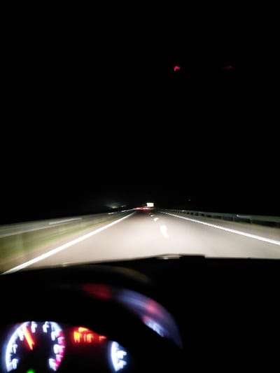 A nighttime view from inside a car driving on a highway. The road is illuminated by headlights, and white lines mark the lanes. A dashboard with gauges, partially visible, shows the car is in motion. Red lights are also apparent on the dash. The surrounding environment is dark, and there is an unfocused blob of red light in the distance.