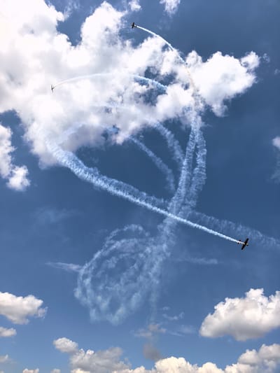 This image depicts an air show with three small airplanes performing aerial maneuvers, leaving behind white smoke trails against a bright blue sky with scattered white clouds. The airplanes are small, single-engine propeller aircraft. One airplane is visible in the upper right quadrant, trailing smoke. Another is in the upper left quadrant, also trailing smoke. A third airplane is in the lower right quadrant, moving diagonally upwards and to the left, leaving a long, distinct smoke trail. The smoke trails form various patterns, including loops and straight lines, suggesting synchronized or individual aerobatic performances. The scene is likely taking place outdoors during daylight, under a clear or partly cloudy sky. The image does not provide any information or visual cues to identify the specific location as Nestiukovo, Russia.