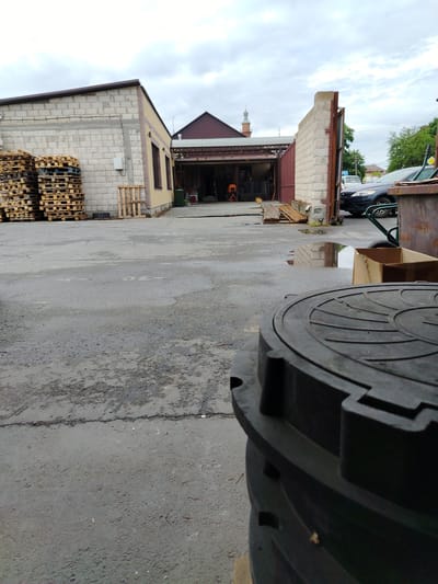 A low-angle, full-shot photograph captures a paved industrial or commercial yard under an overcast sky. In the foreground, a large, dark, circular object, possibly a manhole cover or lid, dominates the lower right quadrant, obscuring the ground. The yard itself is asphalt, wet and showing signs of wear with cracks and patches. To the left, stacks of wooden pallets are piled high against a light-colored brick building. A smaller, single wooden pallet rests near a yellow-bricked section of the building.

Further back, an open-sided structure with a brown roof is visible, appearing to be a workshop or storage area, with various items, including an orange safety cone and equipment, inside. A dark sedan is parked in the background to the right, next to a large, rusty metal container and a hand truck with a wheel. A large puddle reflects the light-colored brick wall of the structure to the right. In the distance, trees and a building with a minaret are visible, suggesting a location within a populated area. The scene appears to be in Beslan, Russia, given the context. There are no people visible in the frame.