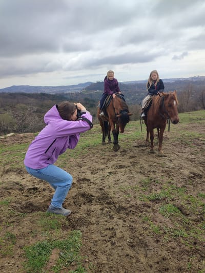 An outdoor scene in Bestuzhevskoye, Russia, depicts three individuals and two horses on a muddy, grassy hillside under an overcast sky. In the foreground, a woman with brown hair, wearing a purple hooded jacket, blue jeans, and gray athletic shoes, crouches low while holding a black DSLR camera to her face, actively photographing the scene ahead. Two young girls, both with blonde hair, are positioned in the middle ground, each mounted on a brown horse. The girl on the left wears a dark purple jacket and dark pants, seated on a dark-maned brown horse with its head lowered. The girl on the right wears a black jacket with pink accents and patterned lower garments, seated on a brown horse with a white blaze on its forehead, also with its head slightly lowered. Both horses are saddled and bridled. The ground beneath them is uneven, a mixture of dark brown mud and sparse green grass, showing hoof prints and shoe marks. In the background, a sprawling cityscape or town is visible nestled among rolling hills and mountains, mostly covered in bare or sparsely foliated trees, under a uniform grey cloud cover.