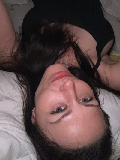 The image shows a close-up, upside-down view of a woman's face and upper body. She has dark hair, fair skin, and is wearing a black top. She is lying on a white, textured surface, possibly a bed or pillow. The lighting is dim, suggesting an indoor setting. There is no indication of the city of Murmansk, Russia, or any specific location. The scene is static, with no observable actions or interactions.